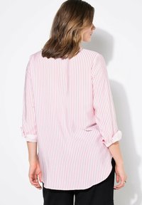 Camisa blanca con rayas verticales rojas, mangas con vuelta, dobladillo redondeado y una textura de tela suave. La vista trasera muestra un diseño minimalista.