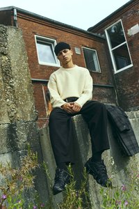 Νέος άντρας με μαύρο σκουφάκι, κρέμα πουλόβερ και μαύρο παντελόνι κάθεται σε σκυρόδεμα τοίχο με πλισέ μπουφάν δίπλα του, με φόντο τούβλινο κτίριο.