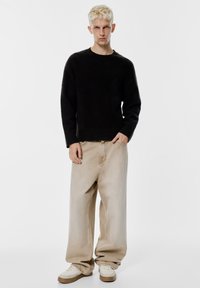 Crni mekani džemper s okruglim izrezom, uparen s bež širokim trapericama. Bijele tenisice s bež detaljima upotpunjuju odjevnu kombinaciju.