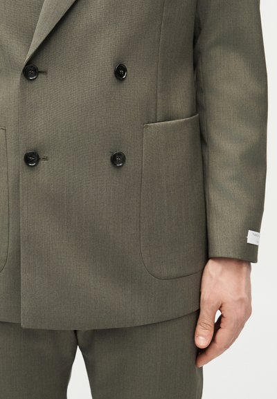 Άνδρας μοντέλο φορά διπλό σακάκι σε ελιά πράσινο με μαύρα κουμπιά και ταιριαστό παντελόνι, με το χέρι χαλαρό δίπλα, μονόχρωμο φόντο.