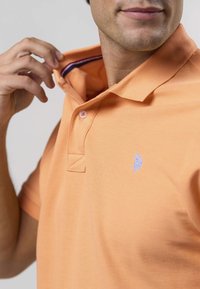 Polo-särk heleoranžist kangast, millel on tekstuuriga viimistlus. Omab kraed, kahte nööpi ja töödeldud sinist logo rinnal.