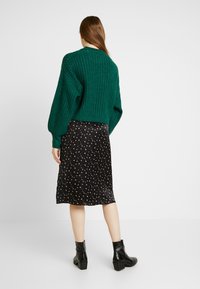 Zelený silný pletený svetr s žebrovanou strukturou, doplněný černou saténovou midi sukní s květinovým vzorem a černými kotníkovými botami.