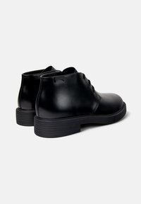 Par de botas de cuero negro hasta el tobillo con cordones, suelas de goma gruesa y un sutil logo bordado en el lado, inclinadas ligeramente hacia atrás.