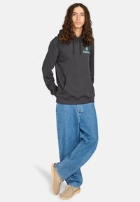 Felpa grigia carbone con tasca a marsupio, cappuccio con cordino e logo turchese sul petto; abbinata a jeans baggy azzurri chiari e scarpe beige.