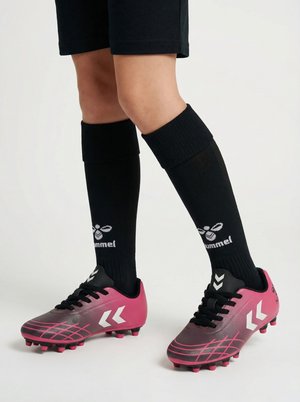 Piernas con calcetines negros Hummel hasta la rodilla y botas de fútbol rosas con cordones negros y logos blancos en forma de chevrón sobre un fondo blanco.