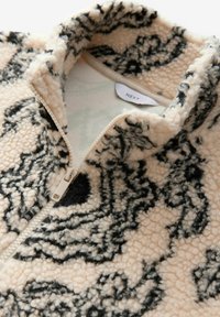 Fleece pullover med høj krave, i en beige bundfarve med sort abstrakt mønster, lynlåslukning og blød tekstur. Mærkets label er synlig.