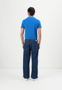 Muškarac okrenut leđima nosi plavu majicu, široke tamnoplave traperice s logom i bijele tenisice na svijetloj jednobojnoj pozadini.