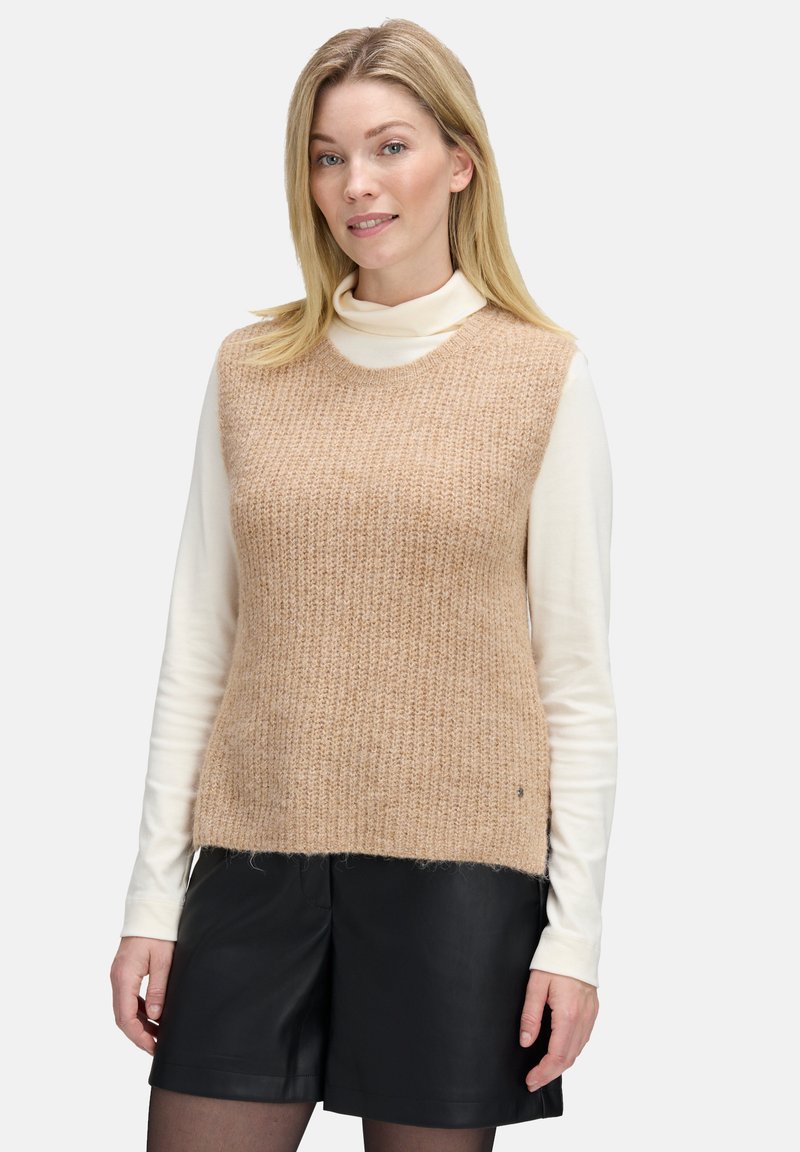 Beżowy sweter bez rękawów z dzianiny o teksturowanym wzorze, noszony na białym golfie, połączony z czarnymi skórzanymi szortami. Prosty dekolt w serek.