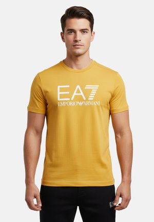 Mann trägt ein senfgelbes EA7 Emporio Armani T-Shirt und schwarze Hose, steht vor einem schlichten hellen Hintergrund.