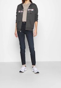 Mörkgrå zip-hoodie med logotyp, beige t-shirt under, mörk denimjeans och vita sportskor med färgglada detaljer.