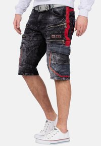Heren cargo shorts van donkerdenim met rode zijaccenten, meerdere ritszakken en een gescheurde textuur. Gecombineerd met witte sneakers.