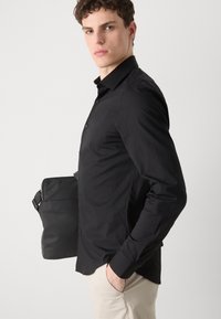 Black button-up shirt with a slim fit, featuring a spread collar and long sleeves. Paired with a black shoulder bag and light beige pants.