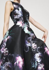 Vestido negro sin mangas con grandes estampados florales en tonos rosa, púrpura y blanco, ajustado en la cintura y con una falda amplia.