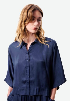 Femme aux cheveux blonds ondulés portant une chemise ample bleu marine à boutons, debout devant un fond clair uni.