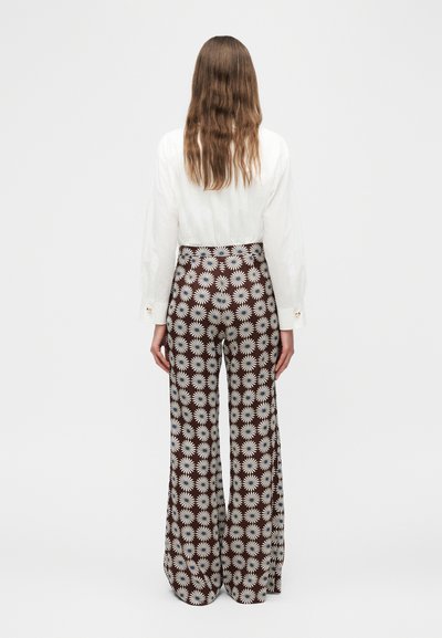 Γυναίκα με μακριά καστανά μαλλιά που φοράει λευκό πουκάμισο και παντελόνι ψηλόμεσο, με φαρδιά μπάτζα και ασπρόμαυρο σχέδιο με μαργαρίτες, φαίνεται από πίσω.