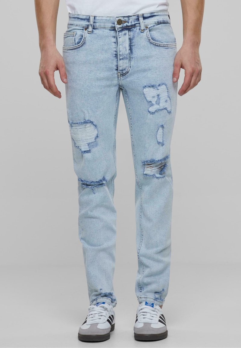 Jean bleu clair déchiré avec plusieurs déchirures, porté avec des baskets blanches et une chemise blanche, montré de la taille aux pieds sur un fond neutre.