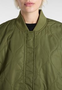 Quilted bomber jacket in olive green, featuring a smooth finish, ribbed collar, and wavy stitching pattern for texture. No visible fastenings.