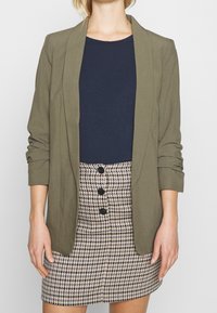 Olivově zelený blazer přes námořnické tričko, kombinovaný s sukní s houndstooth vzorem a černými knoflíky, s ohrnutými rukávy na blazeru.