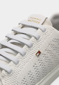 Λευκό πλεκτό αθλητικό παπούτσι με λευκά κορδόνια, μικρό καρφίτσα με το λογότυπο Tommy Hilfiger και κείμενο "Tommy Hilfiger" στην γλώσσα με γράμματα σε χρυσό χρώμα.