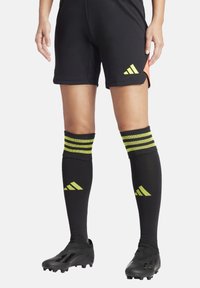 Zwarte voetbalkorte broek met een geel logo, gecombineerd met kniehoge zwarte sokken met drie gele strepen en bijpassende branding. Zwarte voetbalschoenen.