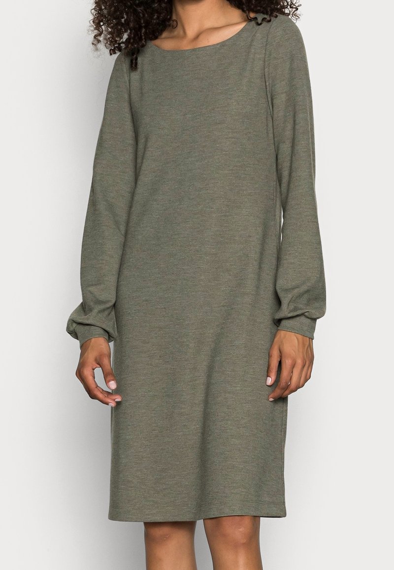 Femme portant une robe olive longue jusqu'aux genoux, à manches longues et col rond, debout devant un fond clair uni.