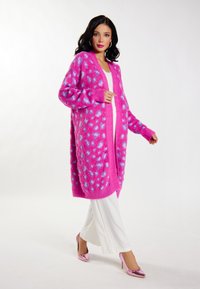Cardigan de gran tamaño en rosa brillante con lunares azules, textura suave, puños acanalados, usado sobre una camiseta blanca y combinado con pantalones blancos y tacones rosas.