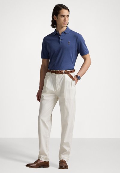 Tricou polo bleumarin cu mâneci scurte și guler, asortat cu pantaloni plisati crem și o curea împletită maro; pantofi din piele maro.