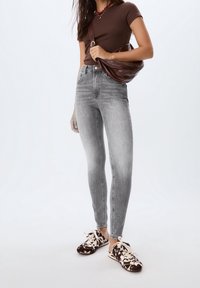 Vaqueros grises de talle alto y ajuste slim, combinados con una camiseta marrón ajustada y zapatillas marrones con estampado, sosteniendo un bolso marrón de hombro.