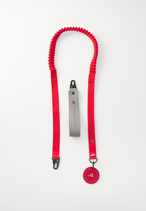 Red elastic leash with a coiled design. Features a gray handle, black clip, and circular tag with brand logo. Durable and flexible material.