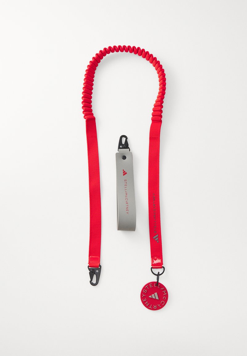 Red elastic leash with a coiled design. Features a gray handle, black clip, and circular tag with brand logo. Durable and flexible material.