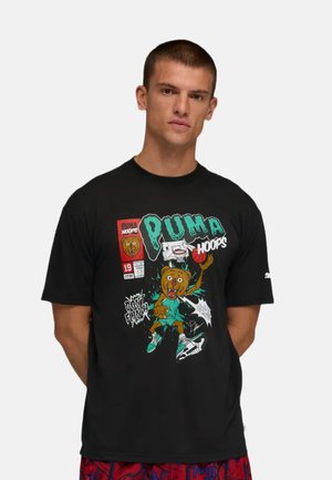 Uomo che indossa una maglietta nera Puma Hoops con un orso dei cartoni animati che gioca a basket e pantaloncini rossi con fantasia, in piedi davanti a uno sfondo chiaro e uniforme.