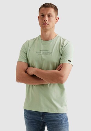 Man met kort haar die een lichtgroen PME Legend T-shirt en een blauwe spijkerbroek draagt, staand met gekruiste armen tegen een effen achtergrond.