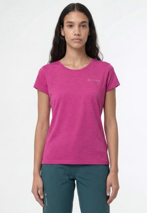 Femme portant un t-shirt à manches courtes unies magenta et un pantalon turquoise, debout devant un fond blanc uni.