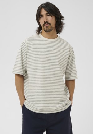 Középhosszú sötét hajú, borostás férfi világosszürke-fehér csíkos rövid ujjú ingben és sötét nadrágban, zsebében a kezével.