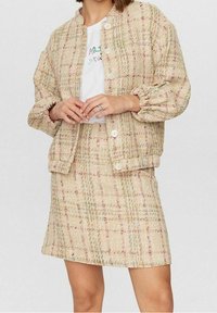 Veste en tweed beige à motif à carreaux pastel, manches bouffantes et gros boutons, assortie à une jupe. T-shirt blanc graphique en dessous.