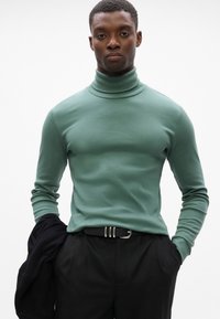 Grüner Rollkragenpullover mit taillierter Passform und langen Ärmeln, kombiniert mit schwarzen Hosen und einem schwarzen Gürtel mit einer silbernen Schnalle.