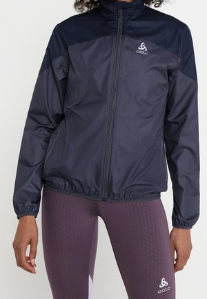 Frau trägt eine marineblaue Jacke mit Reißverschluss und lila strukturierte Leggings, beide mit weißen Odlo-Logos auf der Brust und dem Oberschenkel.