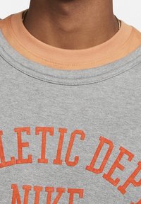 Sudadera gris con letras naranjas que dicen "DEPARTAMENTO ATLÉTICO". Presenta un cuello en capas de color melocotón claro y una tela suave y texturizada.