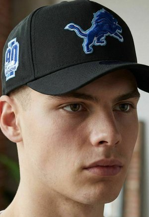 Junger Mann trägt eine schwarze Kappe mit einem blauen Löwenlogo vorne und der weißen Aufschrift "90 Seasons" an der Seite.