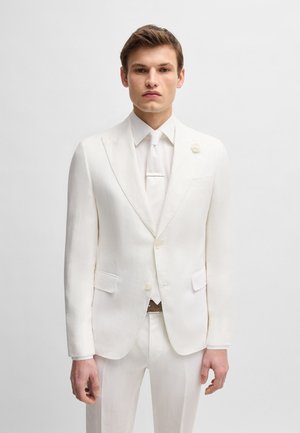Weißer Leinenanzug mit Reversklappen, vorderen Taschen und einem Knopfverschluss. Kombiniert mit einer weißen Krawatte und einem Hemd, das ein minimalistisches Design zeigt.
