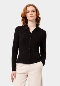 Černá košile s dlouhým rukávem na knoflíky vyrobená z hladké látky s klasickým límečkem, zdobená řadou knoflíků vpředu, doplněná o světle zbarvené kalhoty.