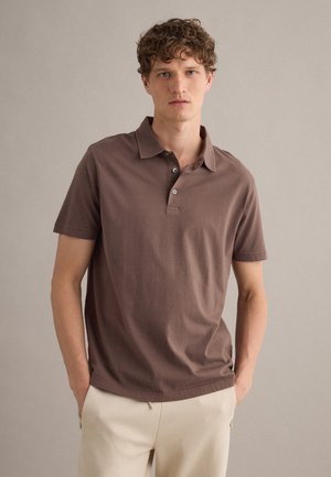 Joven con cabello rizado lleva una camiseta polo marrón de manga corta y pantalones beige, de pie frente a un fondo liso y claro.