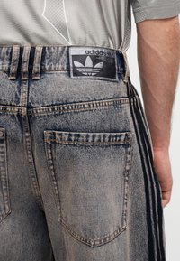 Άτομο που φοράει acid-washed denim σορτς Adidas με μαύρες ρίγες και ορατό λογότυπο στη μέση, μαζί με γκρι πουκάμισο.