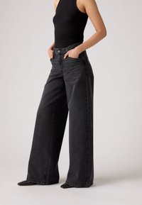 Plati juoda džinsai su aukštu liemeniu, turintys lygią tekstūrą ir klasikinį penkių kišenių dizainą. Sutaikyti su aptemptu juodu viršutiniu drabužiu.