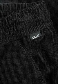 Nahaufnahme von schwarzen, strukturierten Stoffhosen mit elastischem Bund und einem kleinen schwarzen Etikett, auf dem in weißer, geschwungener Schrift "Reell" steht.