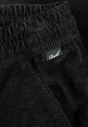 Close-up of black textured fabric pants with elastic waistband and small black label reading "Reell" in white cursive font.