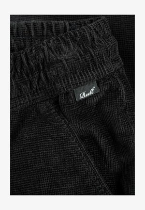 Close-up of black textured fabric pants with elastic waistband and small black label reading "Reell" in white cursive font.