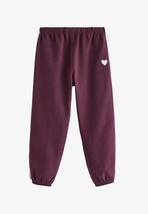 Barn med krøllet hår iført en hvid blomsterlangærmet skjorte, bordeaux joggingbukser med hvide striber og matchende bordeaux sneakers.