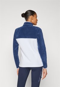 Fleecejakke med blå øvre del og hvit nedre del. Har høy krage og glatt tekstur. Minimalistisk design uten utsmykninger.
