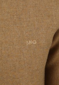 Bruin textiel met een subtiele weving, voorzien van een geborduurd wit "M&G" logo. Het materiaal lijkt zacht en comfortabel te zijn.